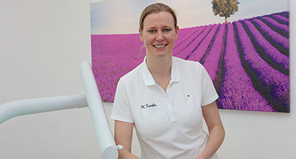 Zahnärztin Kristin Tresko steht im Behandlungszimmer an eine Behandlungseinheit angelehnt, im Hintergrund ein Bild mit einem Lavendelfeld