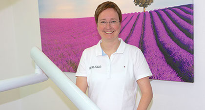 Zahnärztin Dr. Mirja Schulz steht im Behandlungszimmer an eine Behandlungseinheit angelehnt, im Hintergrund ein Bild mit einem Lavendelfeld