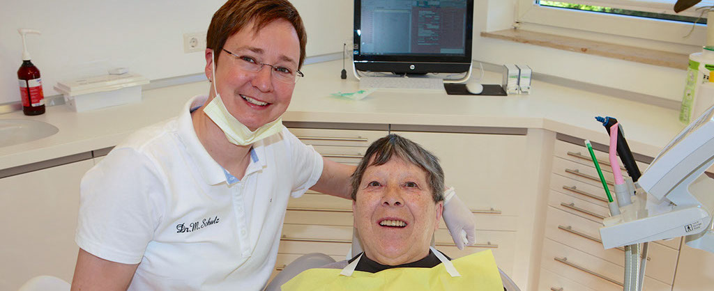 Eine Zahnärztin sitzt im Behandlungszimmer neben einer Seniorin, die lächelnd im Behandlungsstuhl sitzt, im Hintergrund ist Computer zu sehen