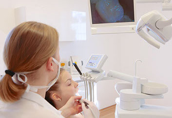 Die Zahnärztin behandelt eine junge Patientin mit Hilfe der intraoralen Kamera, das Bild des Mundraumes ist auf Monitor im Hintergrund zu sehen