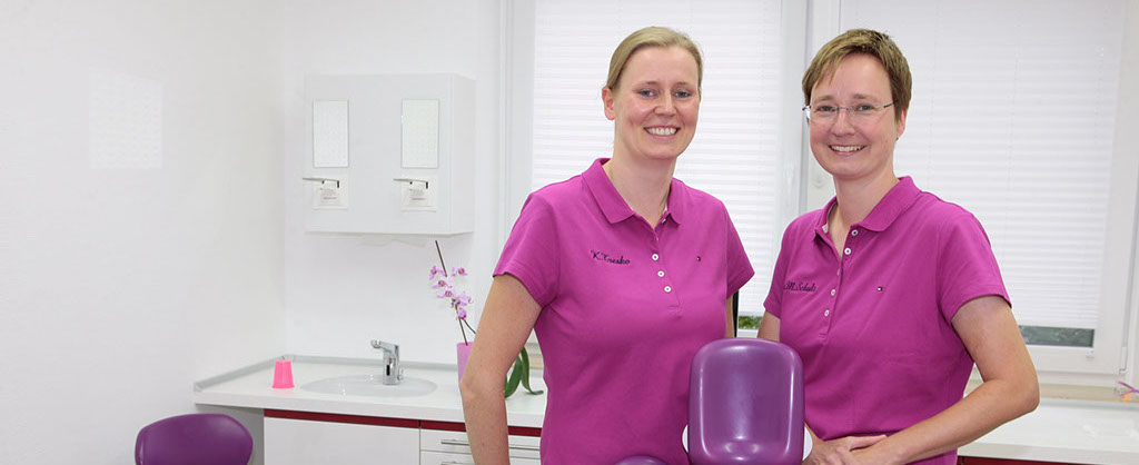 Zwei Frauen stehen in einem Behandlungszimmer hinter einem Behandlungsstuhl, im Hintergrund ist ein Waschbecken zu sehen