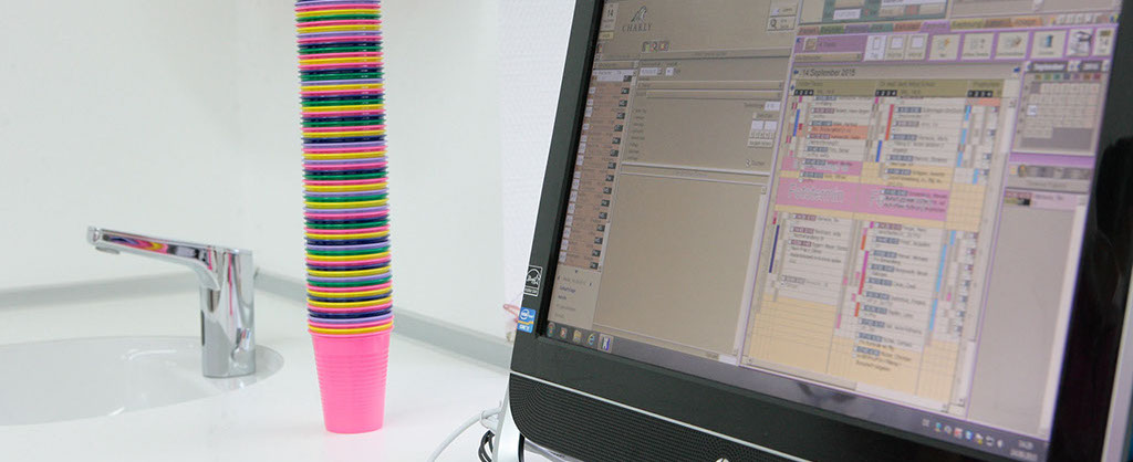 Computerbildschirm mit digitaler Patientenakte im Vordergrund, im Hintergrund stehen bunte Plastikbecher an einem Waschbecken.