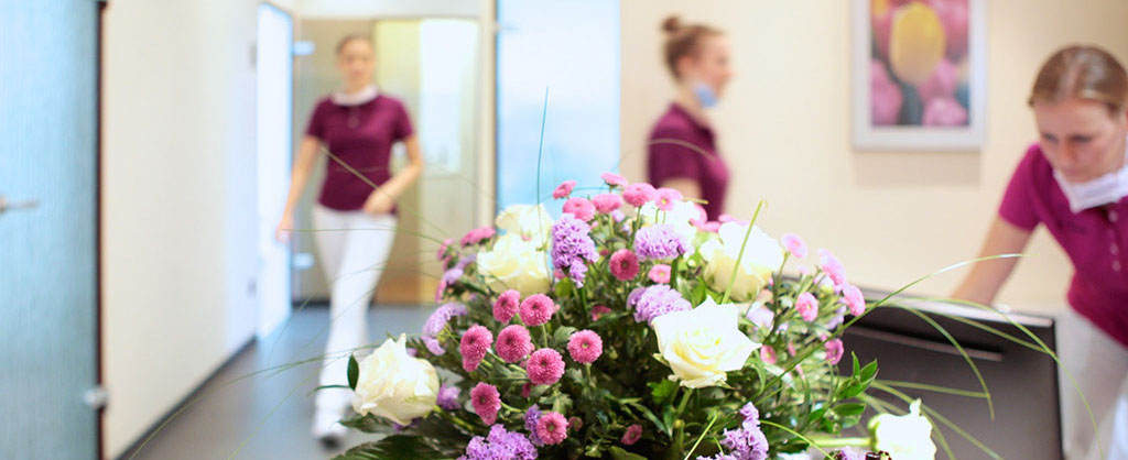 Im Vordergrund ein lilafarbener Blumenstrauß, im Hintergrund drei Mitarbeiterinnen, die in verschiedene Behandlungsräume gehen